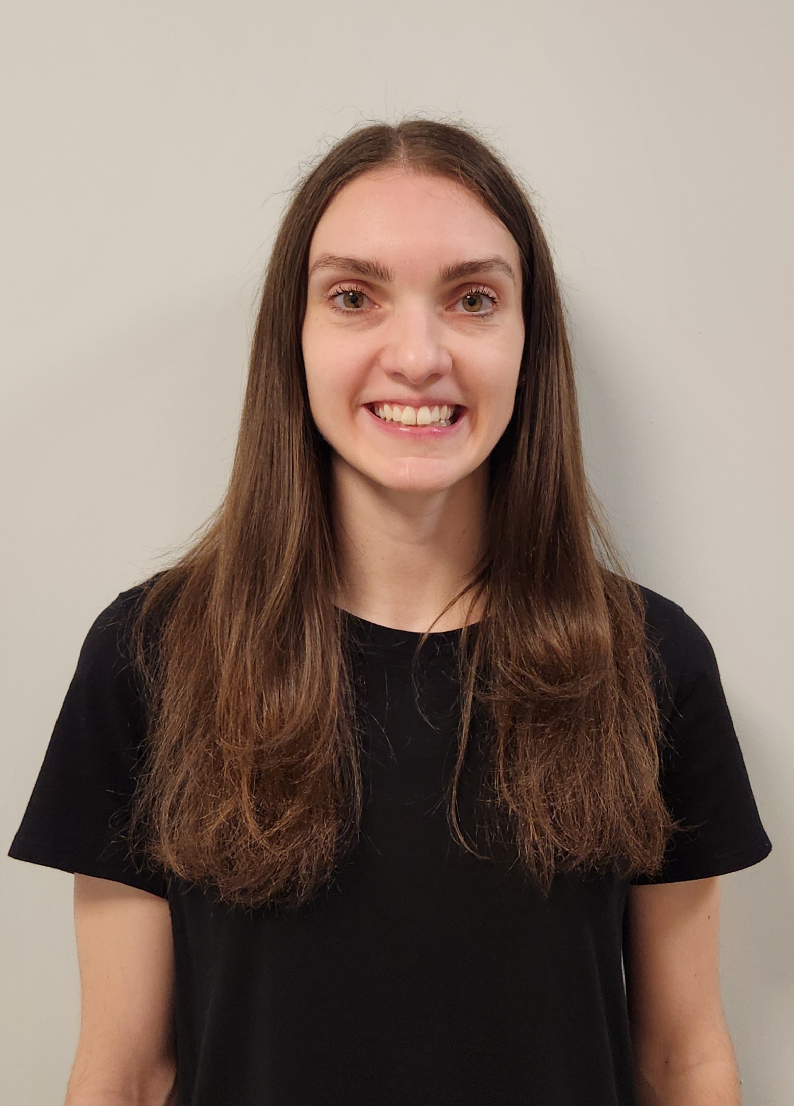 A member of Our Team with straight brown hair and a black shirt stands in front of a plain light-colored wall, smiling at the camera.
