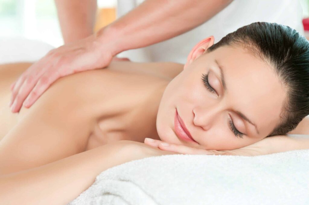 A person with closed eyes lies face down on a massage table while receiving a back massage from another person.
