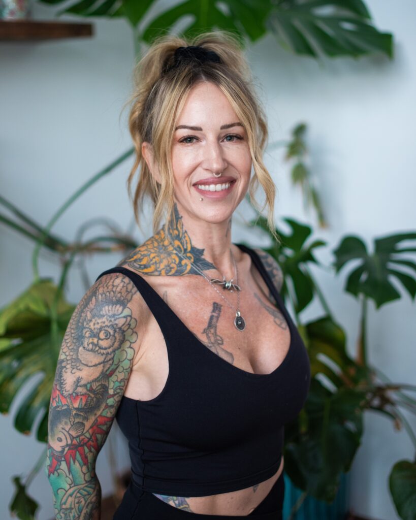 A woman with tattoos on her arm and chest, wearing a black sleeveless top, stands in front of green leafy plants and smiles at the camera—capturing the vibrant energy of Chris Csak Pilates Victoria.