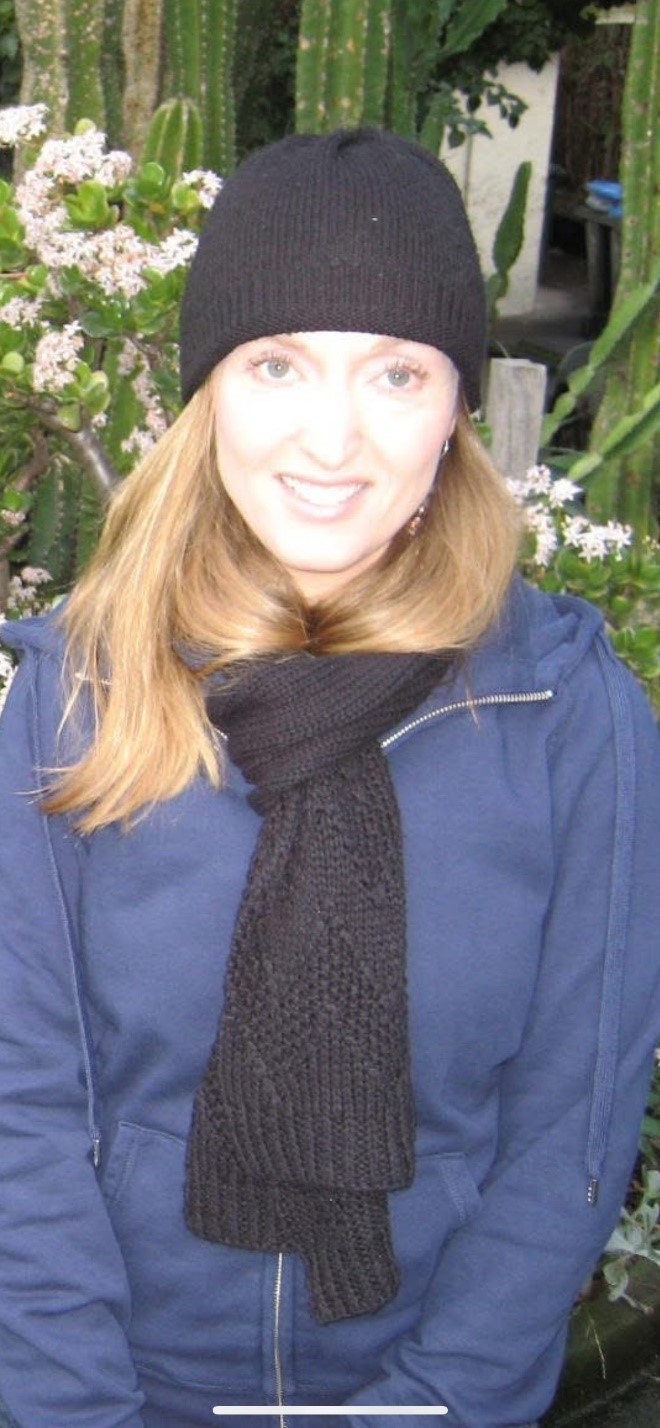 A woman wearing a black knit hat and scarf with a blue jacket stands outdoors in front of green plants and flowers.