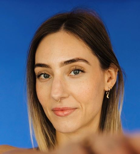 Victoria Pilates instructor Sarah SJ Start, with straight light brown hair and blue eyes, poses in front of a solid blue background. She wears a gold nose ring, small earrings, and has a slight smile.