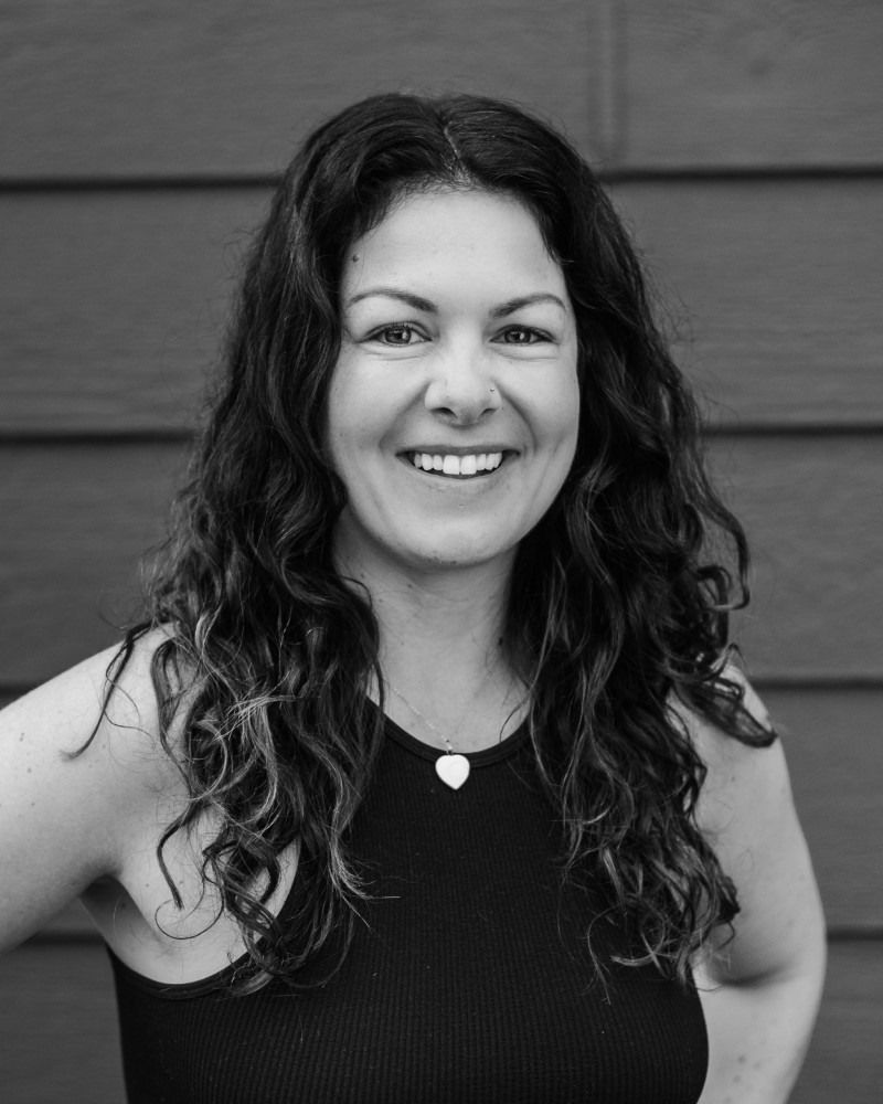 Victoria Pilates instructor Sarah SJ stands smiling in front of a wood-paneled wall, her long curly hair cascading over a sleeveless black top and heart-shaped necklace. Black and white photo.