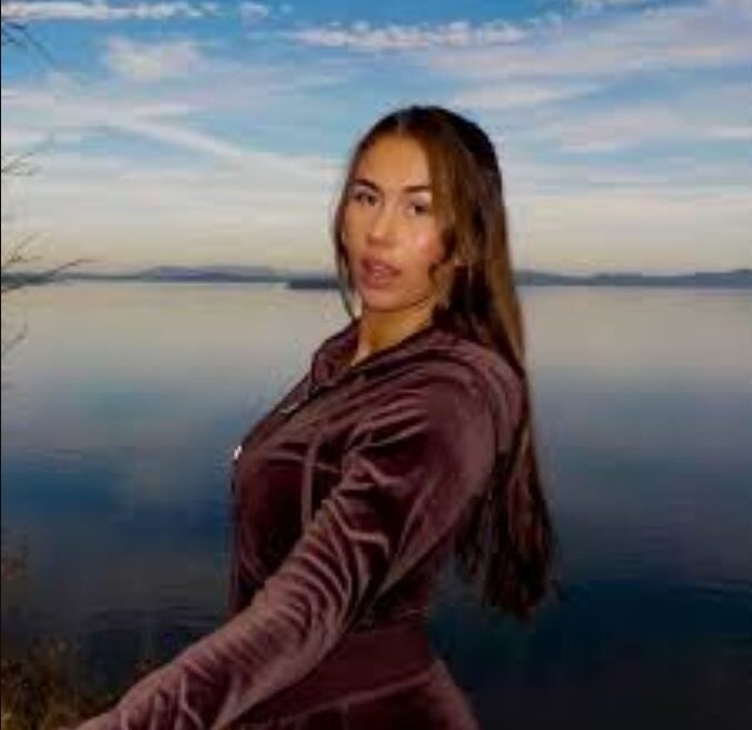 A woman in a brown outfit stands near a calm lake with a blue sky and clouds in the background.