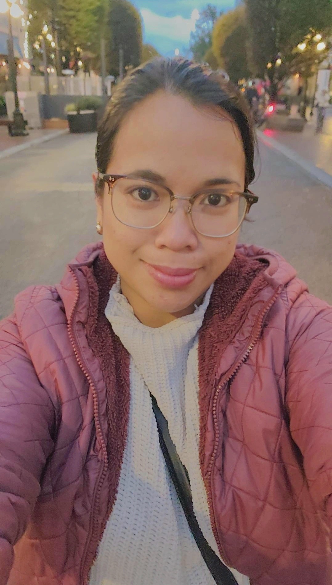A person wearing glasses and a pink jacket takes a selfie on a city street in the evening. Trees and streetlights line the background.